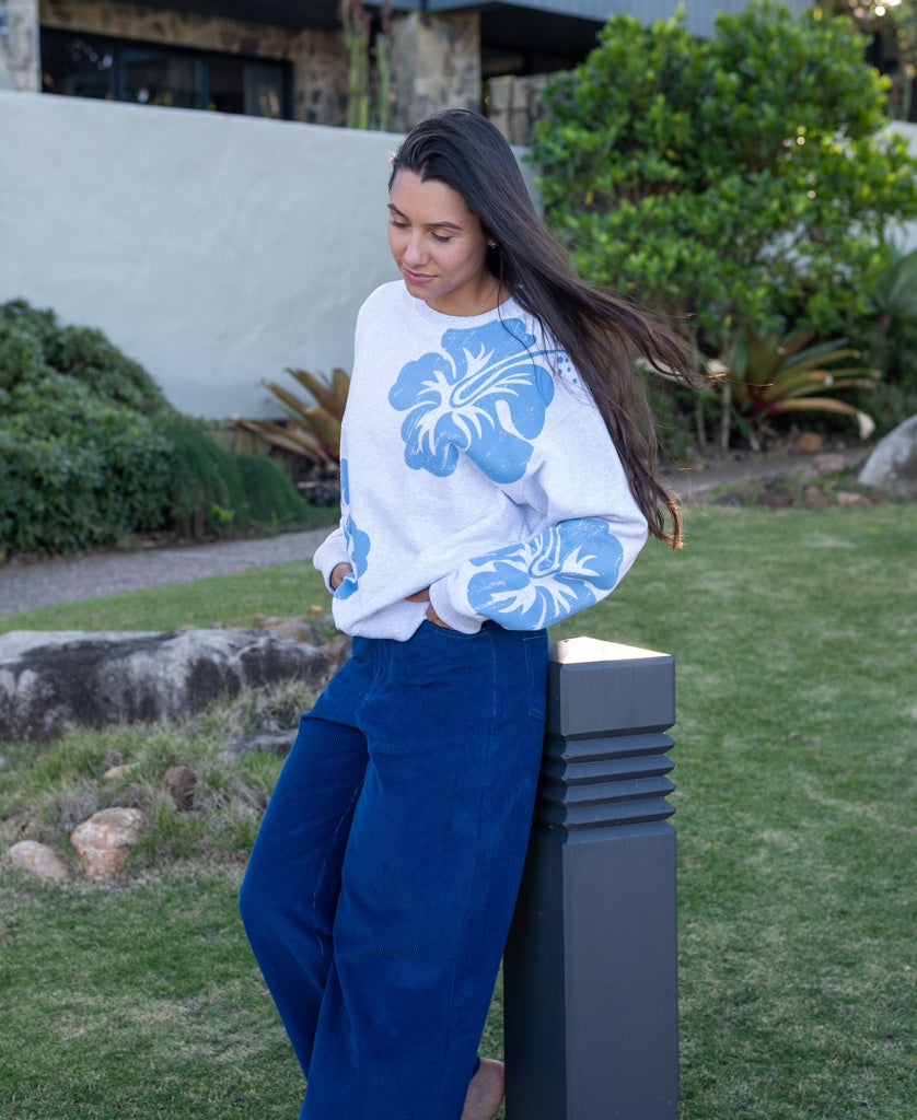 A woman with long dark hair stands on grass, wearing a white sweater with blue floral prints and the Golden Breed Jenny Cord Pant in Navy, styled wide leg, looking down with her hands in her pockets.