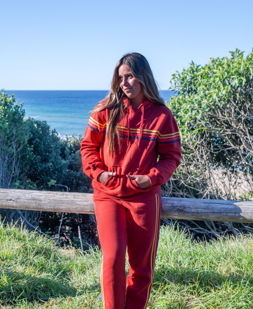 A person in a red Golden Breed Long Island Hoodie stands outdoors by a wooden fence, with greenery and the ocean visible in the background.