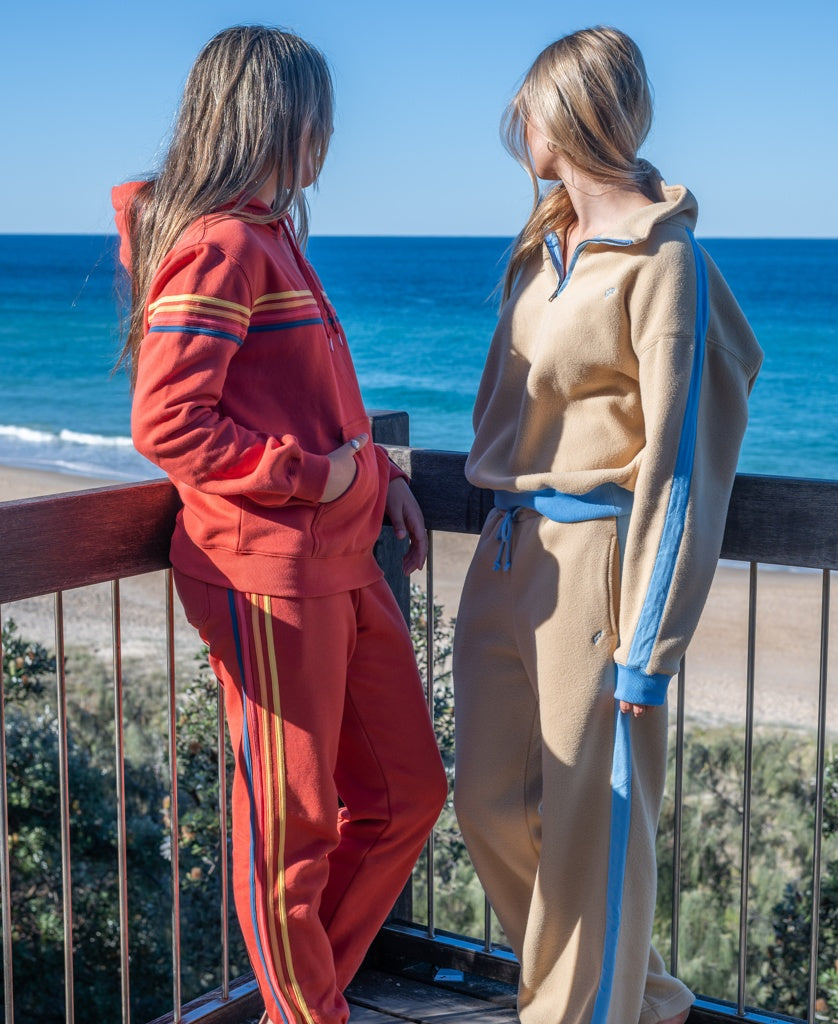 Two people in casual tracksuits, one wearing the Golden Breed Long Island Hoodie | Wash Red, stand on a balcony overlooking the ocean and sandy beach on a clear, sunny day.