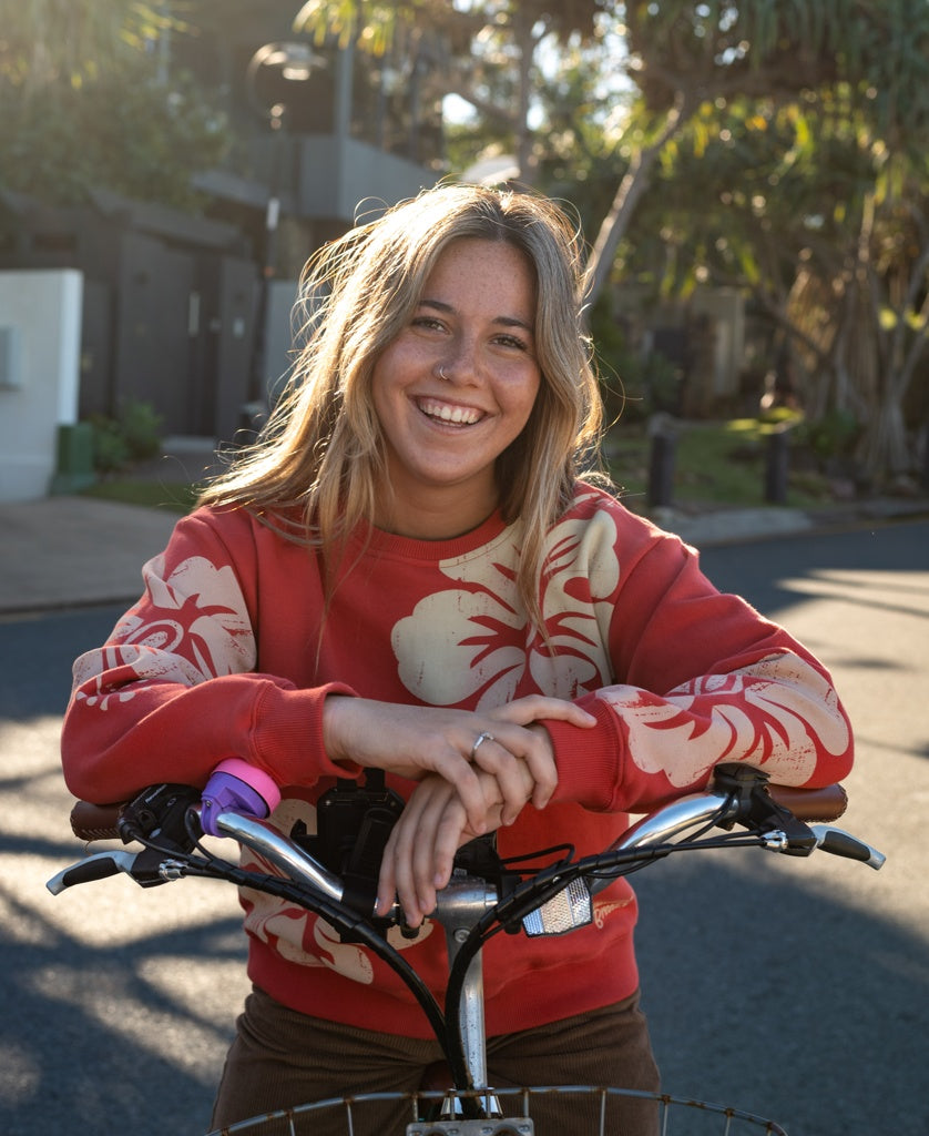 A woman smiles outdoors on a sunny day, resting her arms on a bicycle while wearing the Golden Breed Hibiscus Sweat in Wash Red/Beige, made from 100% cotton fleece.
