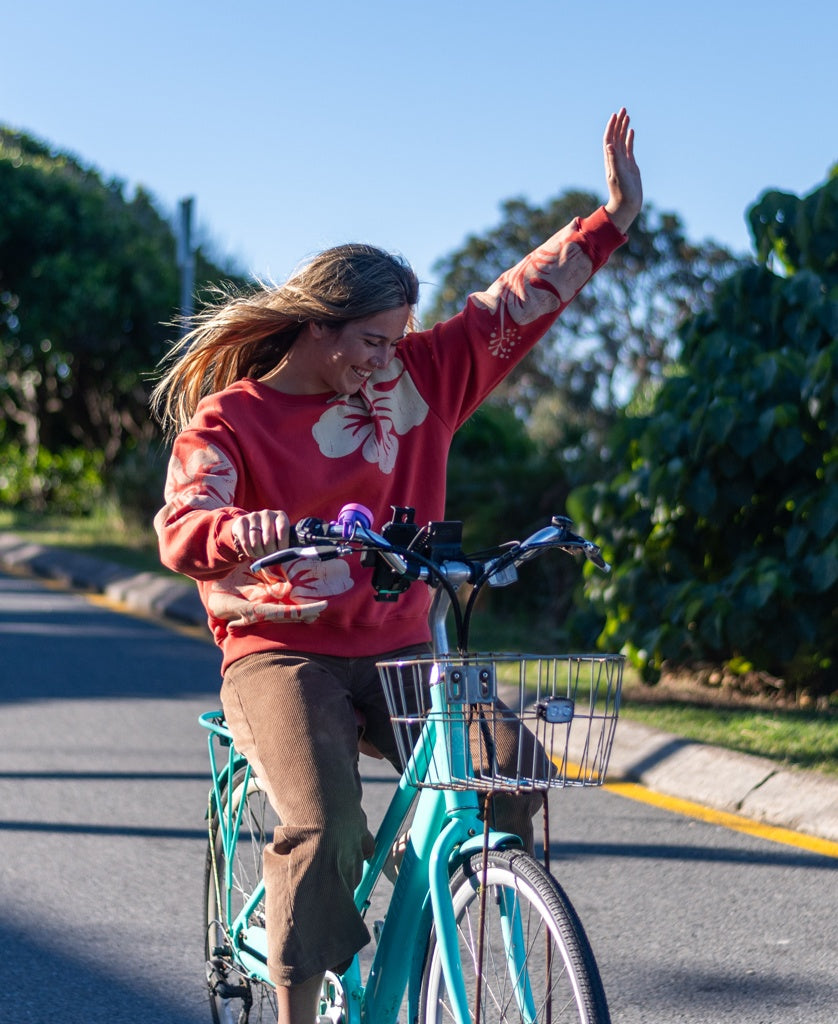 A woman in a Golden Breed Hibiscus Sweat | Wash Red/Beige sweater and brown pants rides a turquoise bike with one hand raised on a sunny, green-lined road.