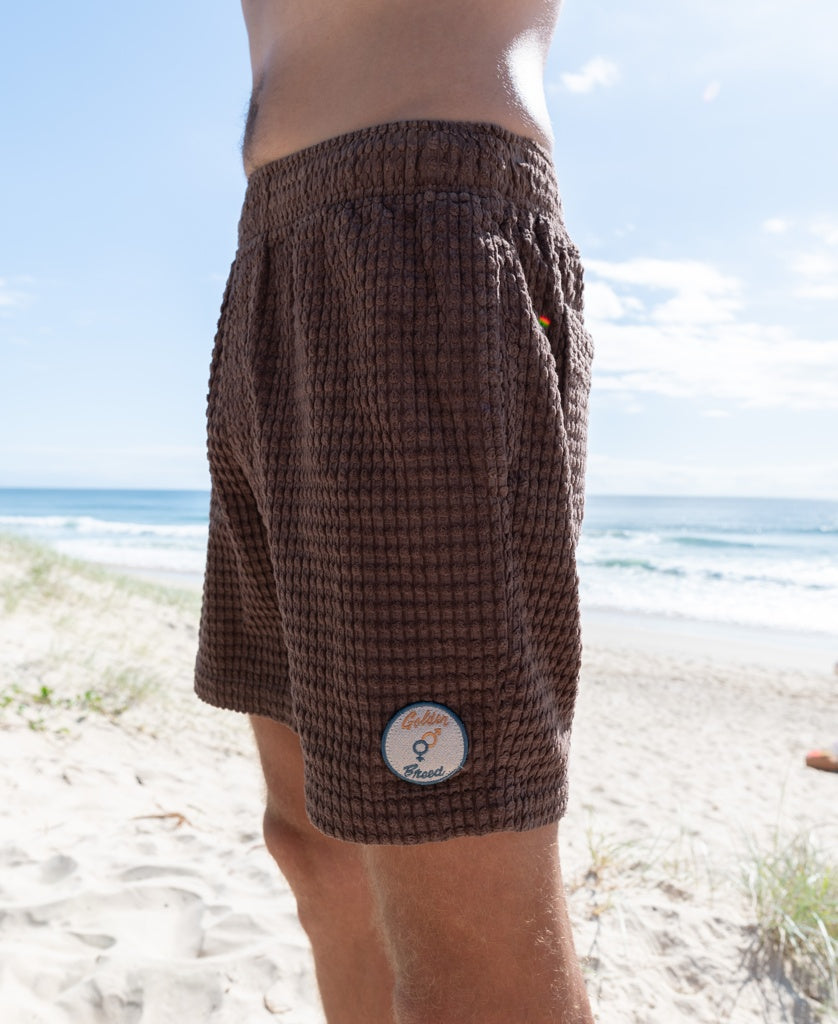 A person wearing Golden Breeds El Shorto Walkshort in Wash Brown, featuring a small round patch and elastic waist, stands on a sandy beach with the ocean and blue sky behind them.