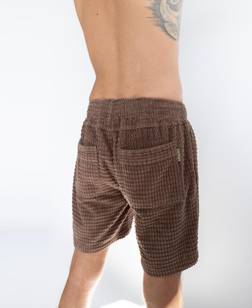A person shown from the back wears Golden Breeds El Shorto Walkshort in Wash Brown—comfortable, textured shorts with an elastic waist cord, highlighting a tattoo on their upper left back.