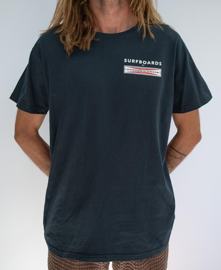 Person with long hair stands against a plain background wearing a navy Gordon & Smith G&S Corp Logo Tee and patterned shorts, with arms behind their back.