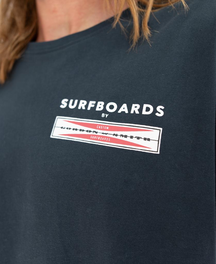 Close-up of a person wearing the G&S Corp Logo Tee | Navy by Gordon & Smith, featuring SURFBOARDS and a red and white graphic on the chest—ideal for fans of classic surf apparel.
