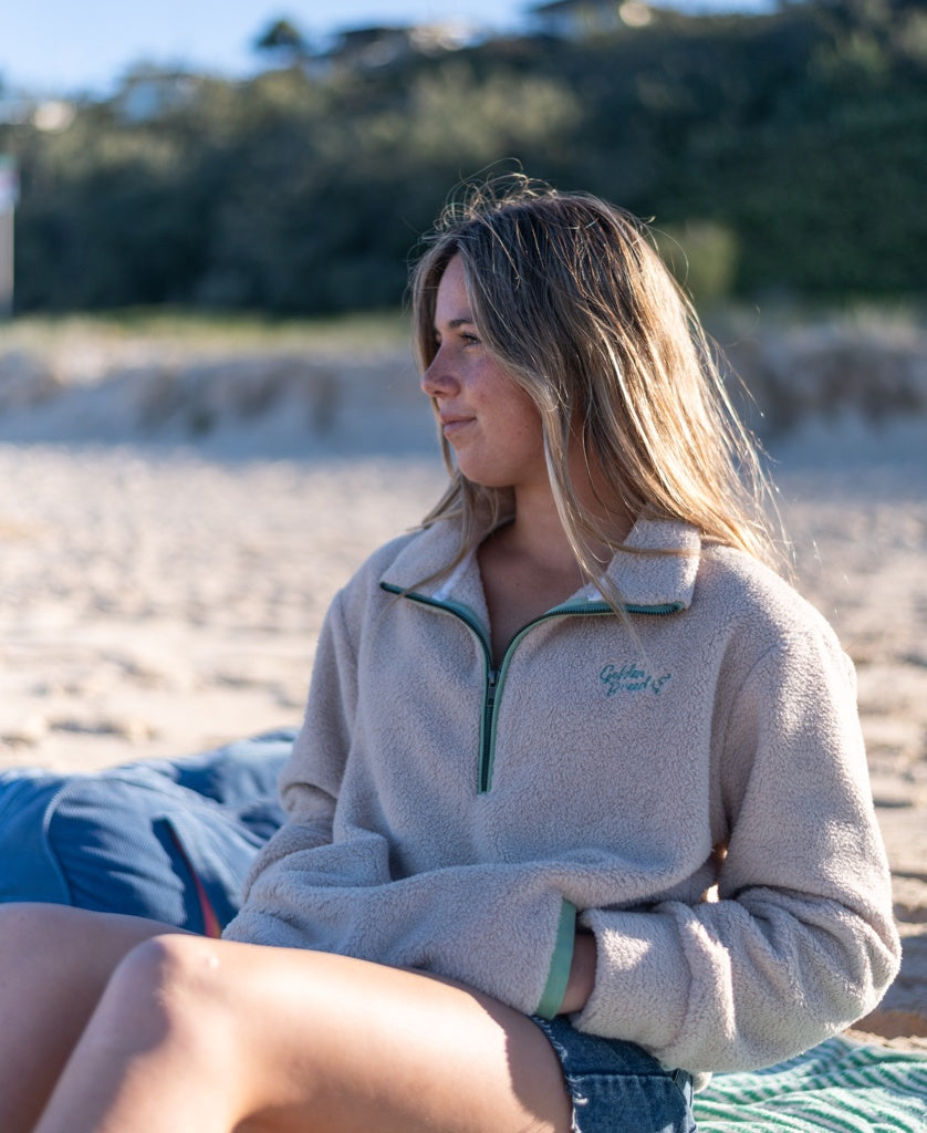 A young woman in the Golden Breed Sammie 1/2 Zip | Beige sits on a beach towel, gazing sideways with sand and greenery in the background.