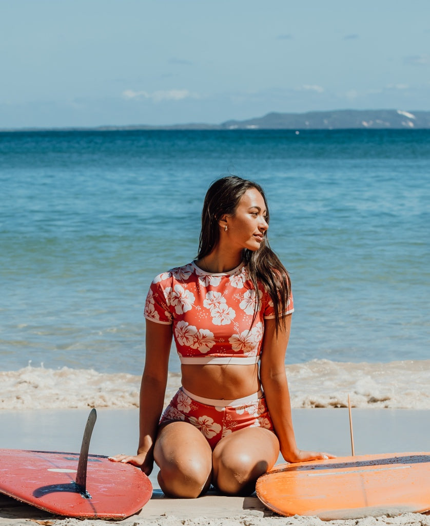 A woman wears the Golden Breed Indie Crop Tee in Hibiscus Wash Red / Beige, sitting on sand between two surfboards at the beach with the ocean and distant land in view.