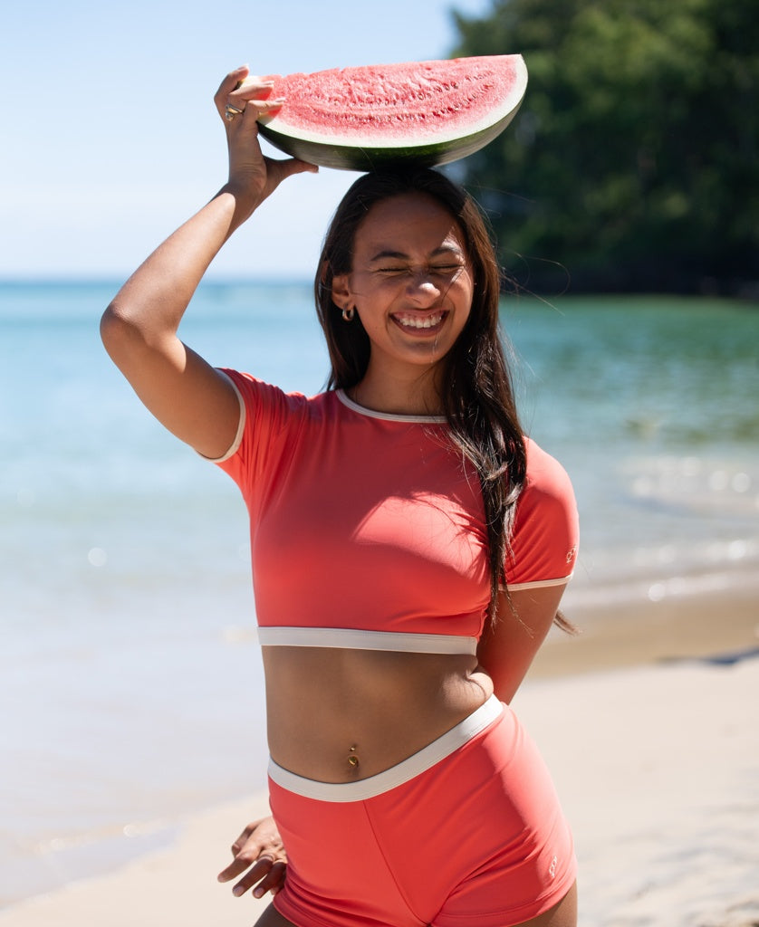 A woman stands on the beach, smiling and lifting a watermelon slice above her head, wearing Golden Breeds Indie Crop Tee Retro in Surf Red / Off White.