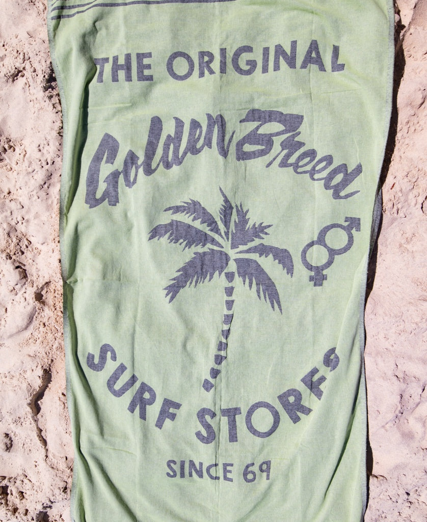 The GB Otto Towel | Green by Golden Breed is a soft Turkish cotton towel featuring The Original Golden Breed Surf Stores Since 69 and a palm tree print, shown lying on sand.
