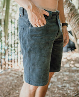 A person wears Golden Breeds Floyd Cord Walkshort in Wash Black, made from 100% cotton corduroy with a structured waist, standing outdoors with one hand in their pocket amid trees and sunlight.