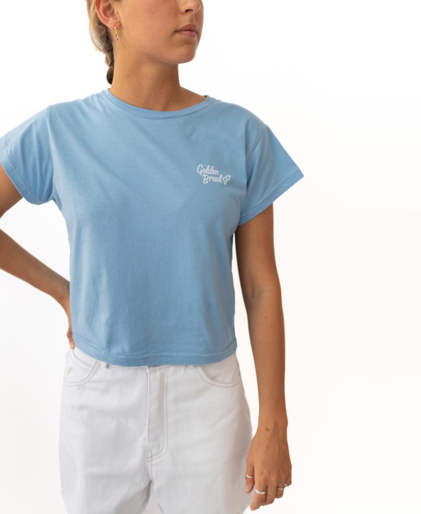 A person wearing the Hattie Tee in Azure by Golden Breed, a light blue cropped top with a vintage Golden Breed chest print, paired with high-waisted white pants and posing against a plain white background.