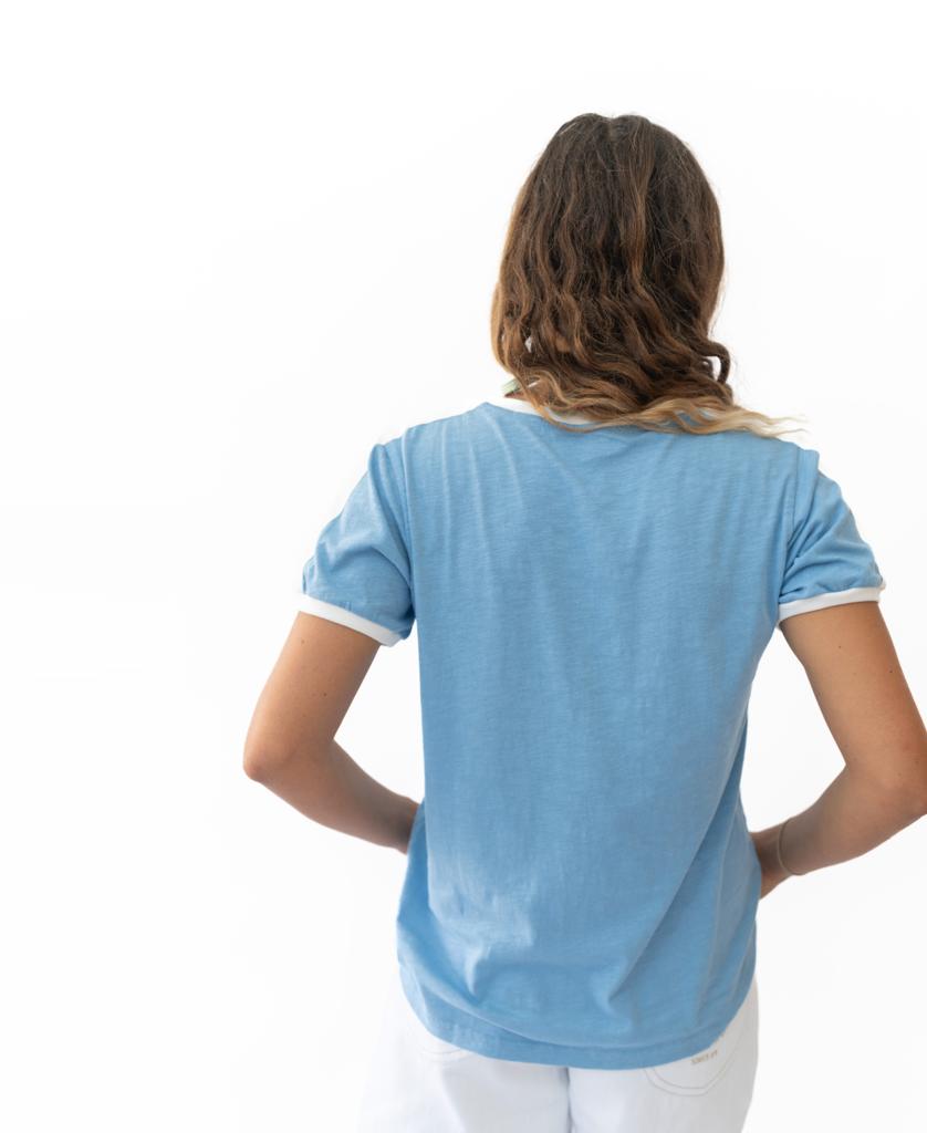 A person with wavy brown hair stands facing away from the camera, wearing Golden Breed’s Ringer Tee in Azure and white pants against a plain white background.