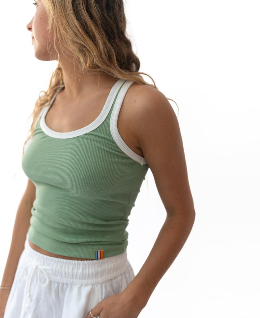 A woman with wavy hair wears the Golden Breed Flick Tank in Moss and white pants, standing against a plain white background and looking to the side.