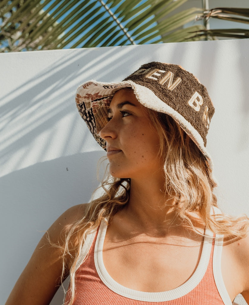 A woman in a tank top wears the Golden Breed Ferry Terry Bucket Hat in Wash Brown, standing against a white wall with palm leaf shadows and looking to the side.