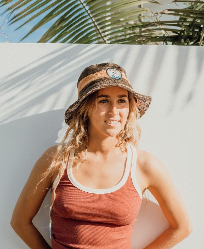 A woman wearing the Golden Breed Nathan Terry Bucket Hat in Wash Brown and a brown tank top stands in sunlight against a white wall, with palm leaf shadows behind her.
