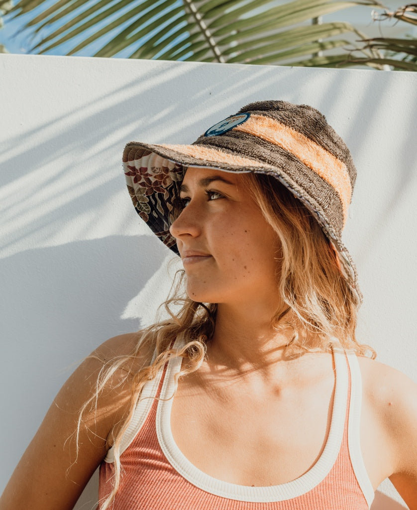 A woman wearing the Golden Breed Nathan Terry Bucket Hat in Wash Brown and a tank top stands in sunlight as palm leaf shadows fall on the white wall behind her.