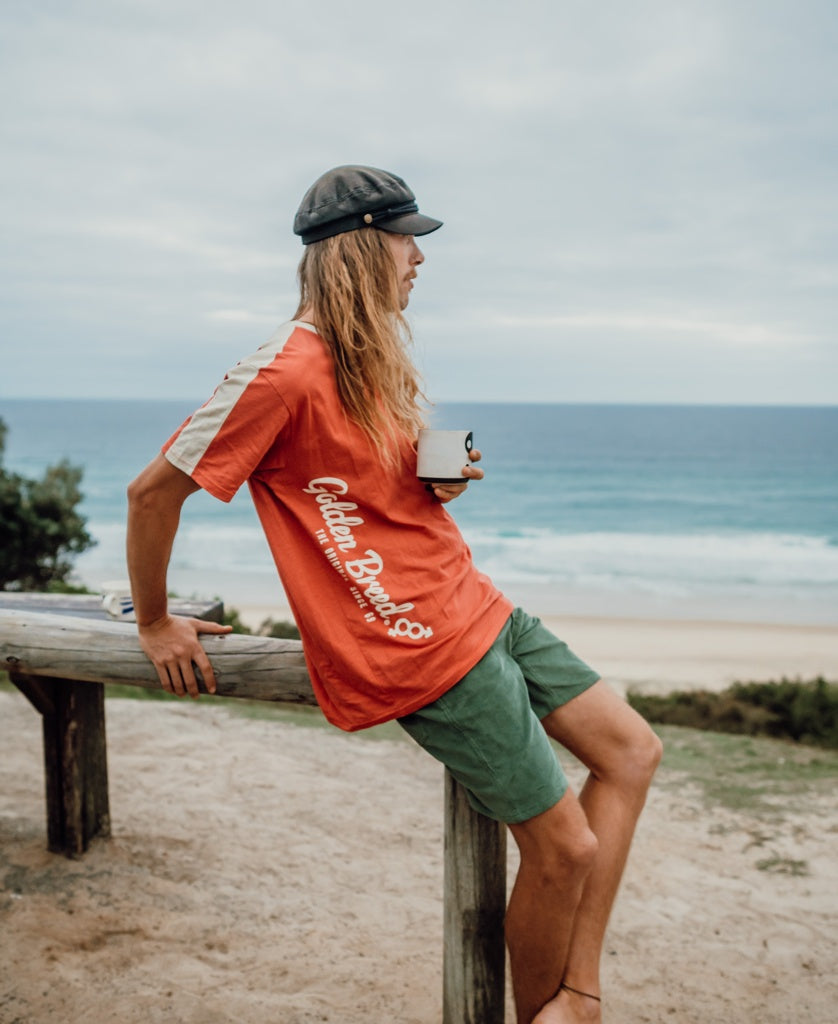 A person with long hair in a cap, Golden Breeds Mitch Tee | Wash Red, and green shorts sits on a wooden rail holding a mug, gazing at the beach and ocean.