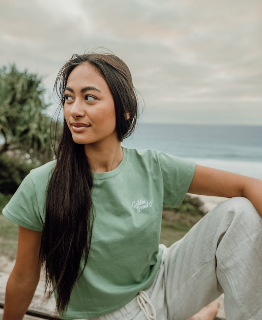 A woman with long dark hair wears the Golden Breed Hattie Tee in Moss and light pants as she sits outdoors near the ocean, looking to the side.