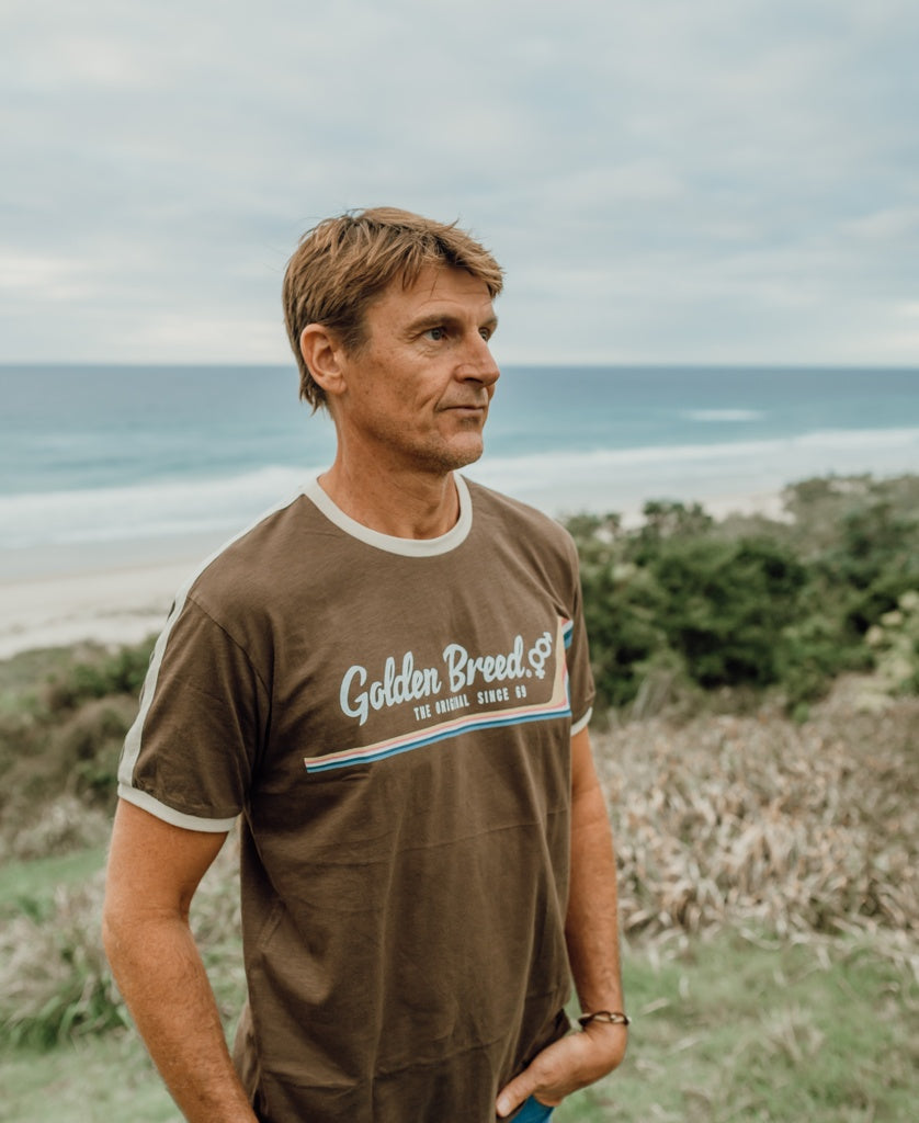 A man in the Golden Breed Retro Ringer Tee in Wash Brown stands by a grassy area near the ocean under a cloudy sky, channeling relaxed 70s vibes.