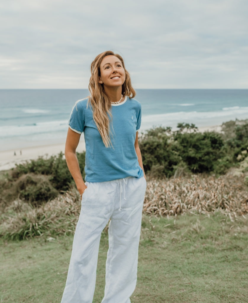 A woman stands on grass by the beach, wearing the Golden Breed Ringer Tee | Azure and white pants, with the ocean and a cloudy sky in the background.