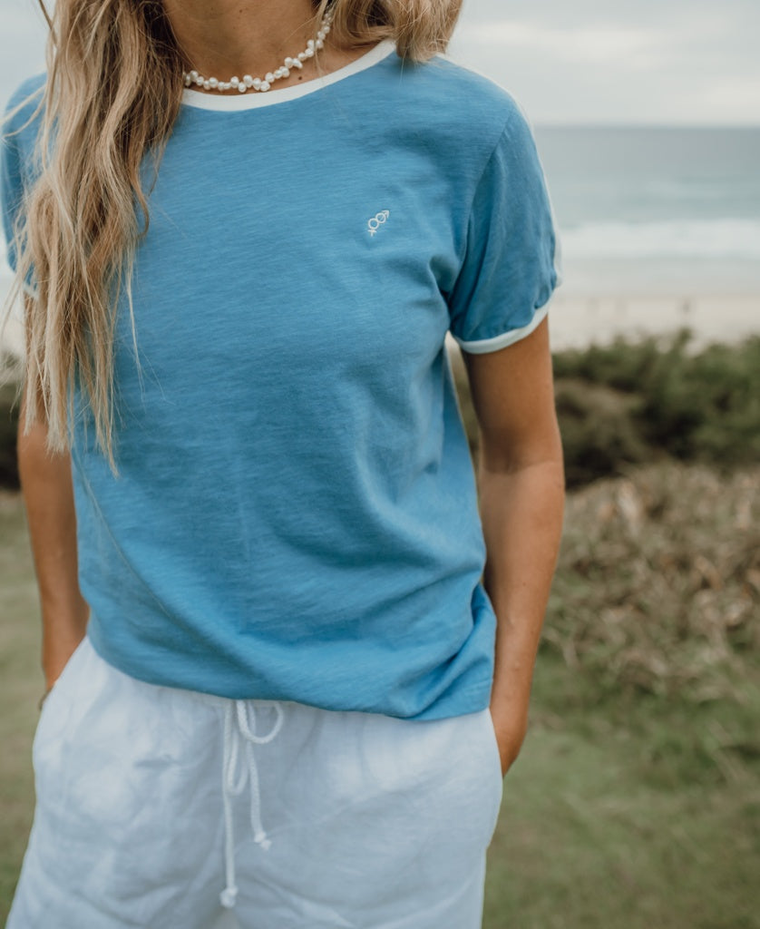 A person wearing the Golden Breed Ringer Tee | Azure and white shorts stands outdoors with hands in pockets, beach and ocean in the background.