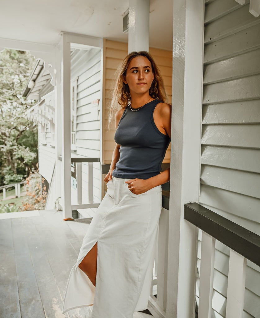A woman in a navy tank top and the Golden Breed Nellie Maxi in Off White leans against a porch railing, gazing into the distance.