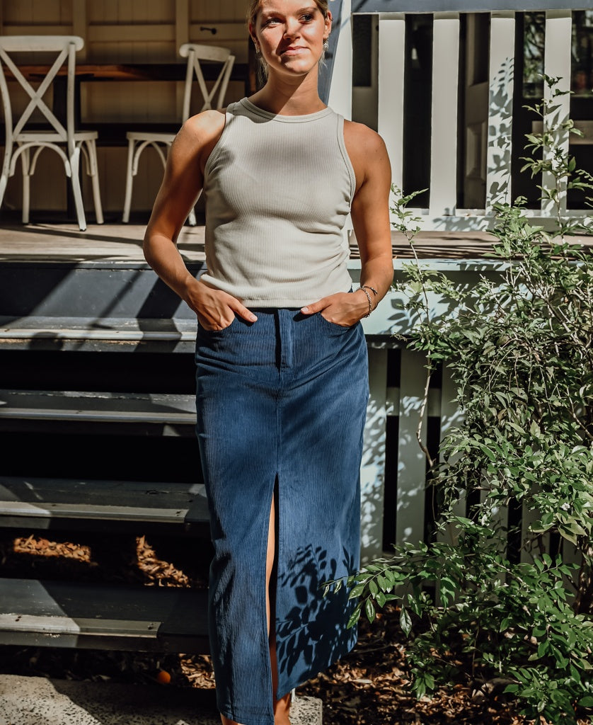 A person stands outdoors in sunlight, wearing a white sleeveless top and the Golden Breed Nellie Maxi Skirt | Petrol, featuring a front slit and pockets, near a shaded porch with green plants.