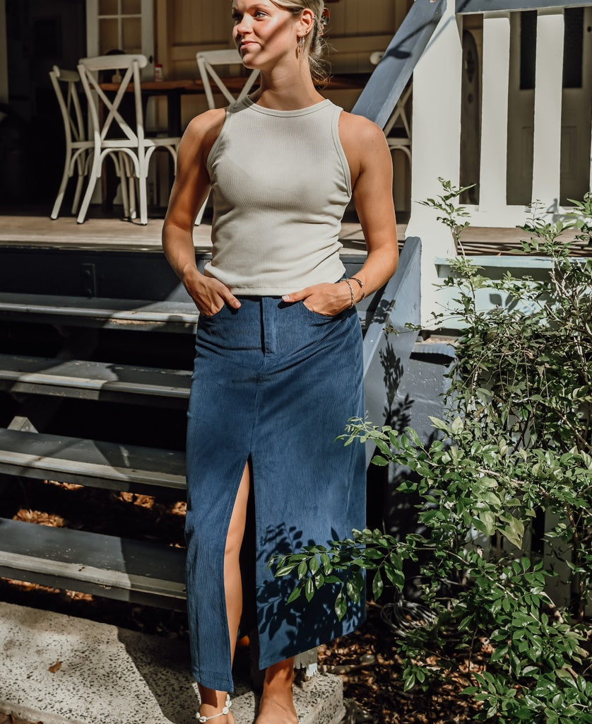 A woman stands outdoors near steps, wearing a sleeveless beige top and the Golden Breed Nellie Maxi Skirt in Petrol with a front slit, her hands tucked in the pockets.