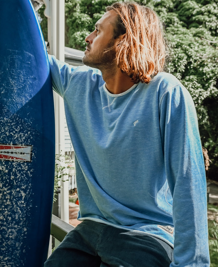 A man with shoulder-length hair wears the Golden Breed Classic Rag Top | Sky Marle as he sits near a surfboard, channeling retro surf style and gazing to the side against a backdrop of greenery.