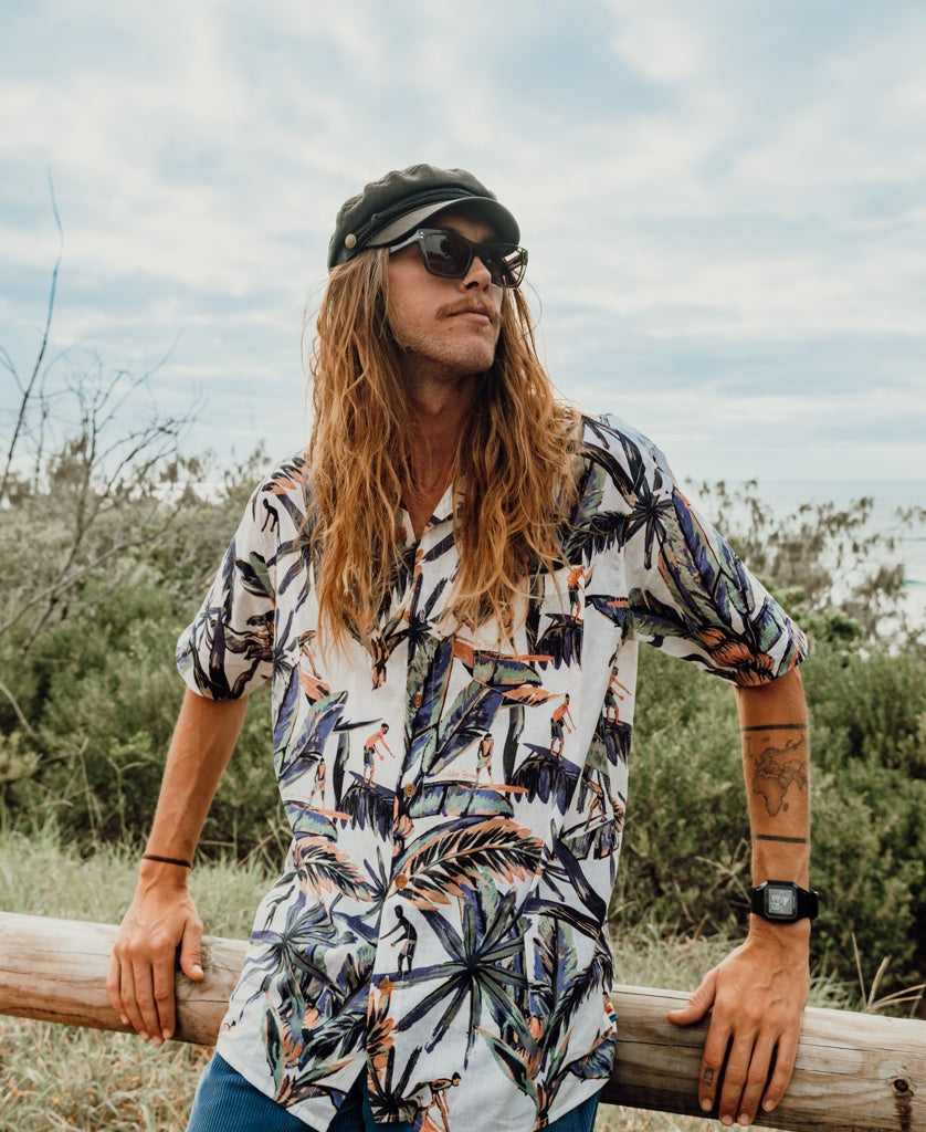 A man with long hair and sunglasses, wearing a black cap and the Golden Breed Surfer Palm Shirt in Off White, stands outdoors by a wooden fence surrounded by greenery under a cloudy sky.