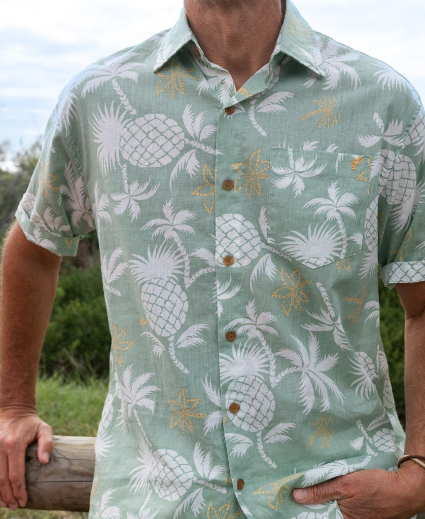A man in the Golden Breed Pine-Palm Shirt | Sage, featuring a light green cotton linen fabric with a pine palm print and oversized relaxed fit, stands outdoors, resting one hand on a wooden rail.