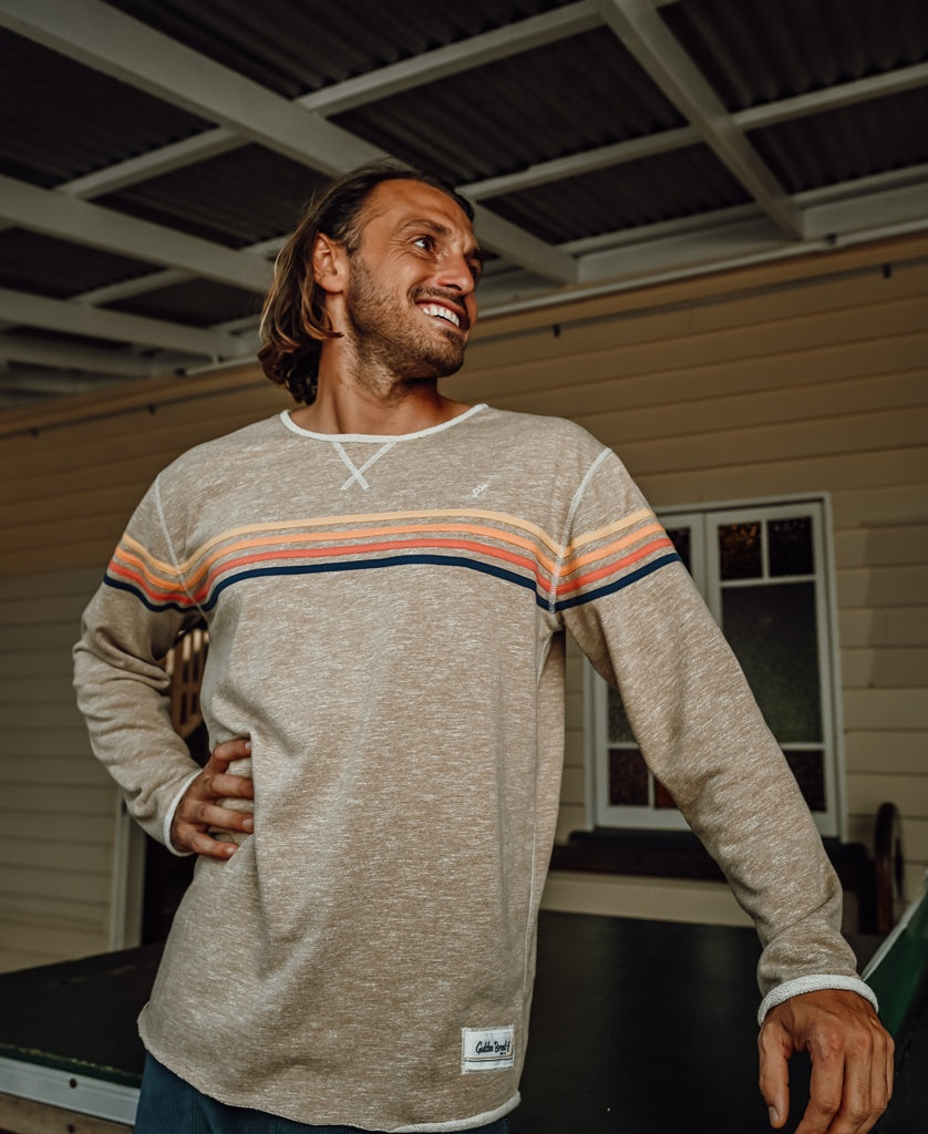 A man with long hair stands indoors, smiling, wearing the Golden Breed Long Island Rag Top in Coffee Marle, featuring colorful horizontal stripes for a retro style look.