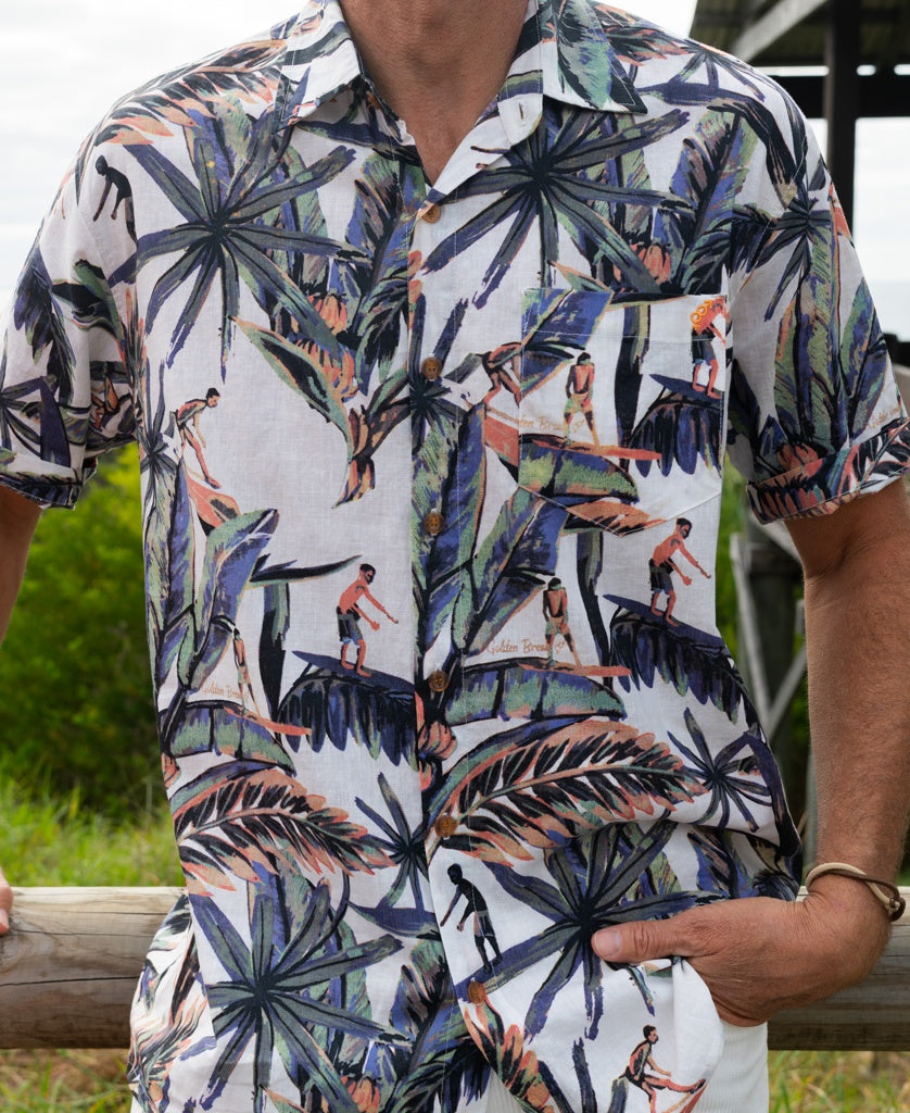 A man in the Golden Breed Surfer Palm Shirt | Off White, featuring a colorful tropical and surfing-themed print, stands outdoors with one hand resting on a wooden rail.