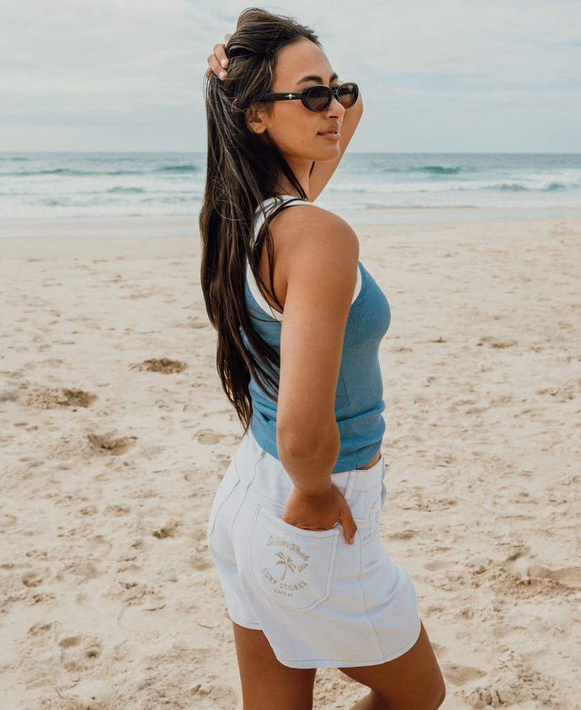 A woman in sunglasses, a blue tank top, and Golden Breed’s Rudi Short in Off White Denim stands on a sandy beach, one hand in her pocket, facing sideways toward the ocean.