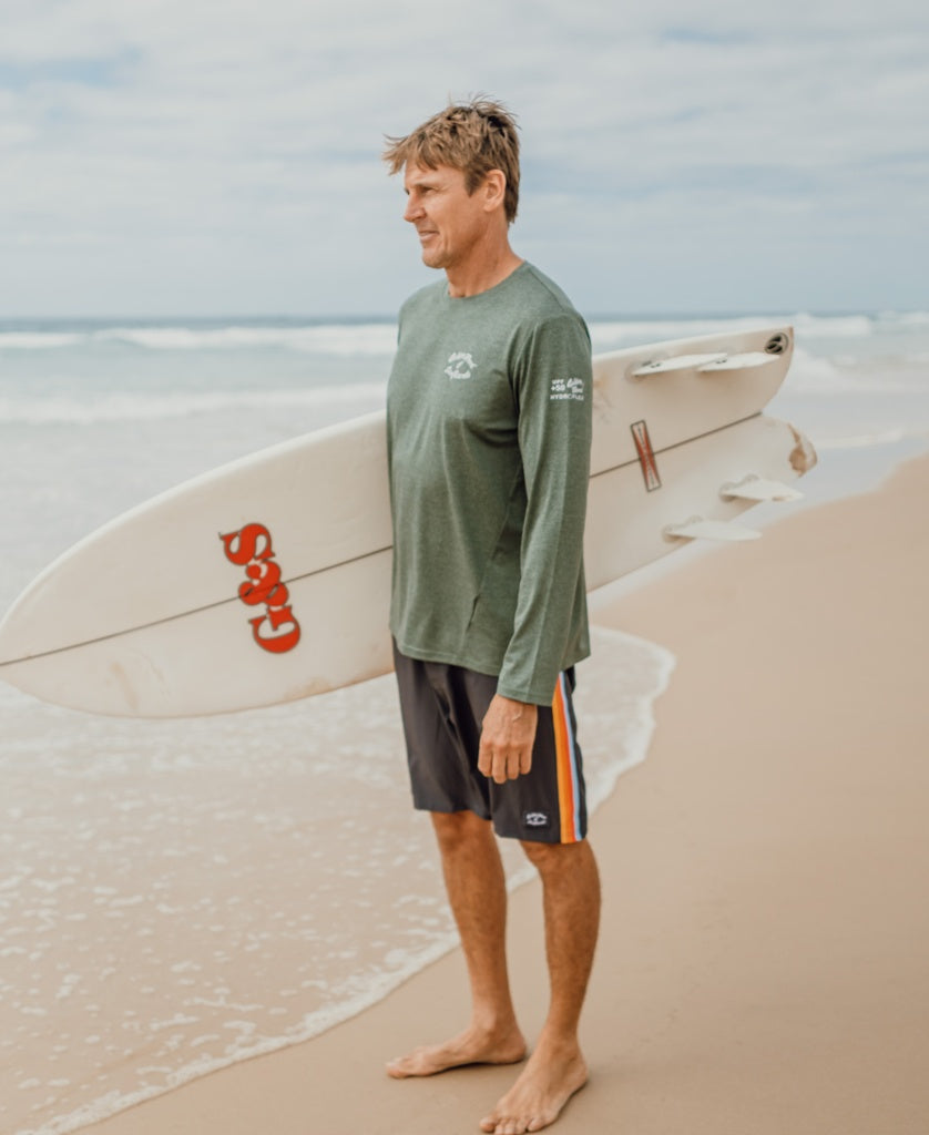 A man stands barefoot on the beach, wearing a Golden Breed Pebbles L/S Rash Shirt in Green Marle with 50+ UPF sun protection, holding a white surfboard under his arm and facing the ocean beneath overcast skies.