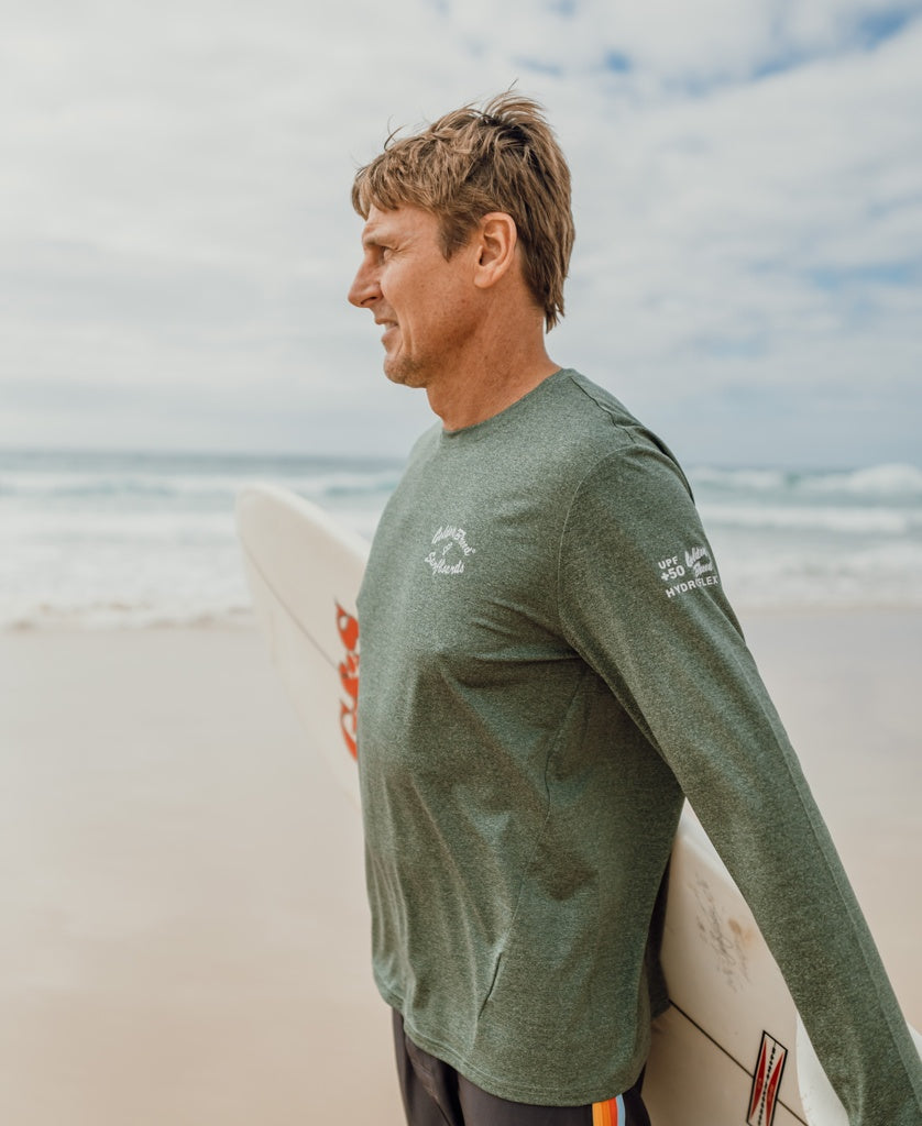 A man stands on the beach holding a surfboard, wearing Golden Breeds Pebbles L/S Rash Shirt in Green Marle for 50+ UPF sun protection and shorts, gazing at the ocean.