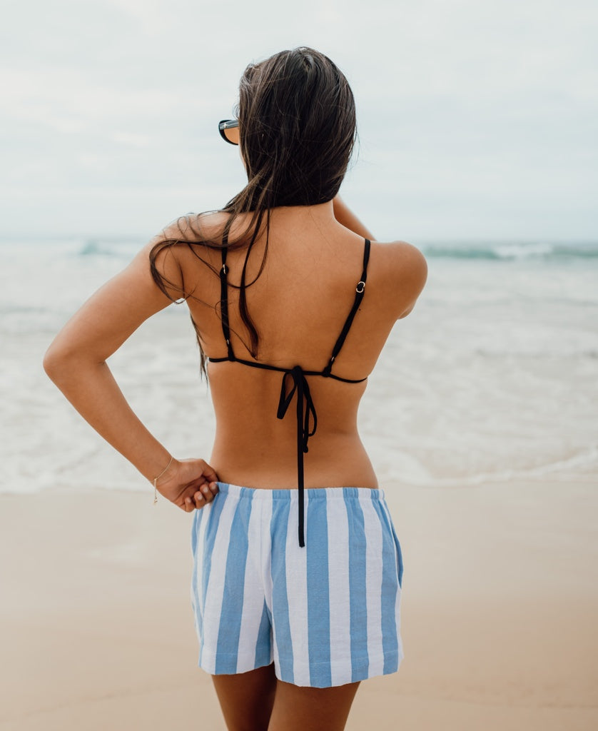 A woman in sunglasses wears Golden Breed’s Molly Short in Azure/White, standing with her back to the camera on a sandy beach—showcasing this summer essential in breezy cotton.