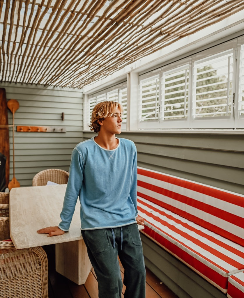 A young person in the Golden Breed Classic Rag Top in Sky Marle leans on a table in a sunlit patio with striped benches and wicker chairs.
