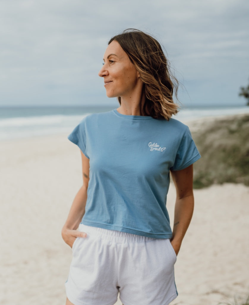 A woman stands on a sandy beach, wearing the Golden Breed Hattie Tee in Azure and white shorts, gazing to the side with the ocean and cloudy sky behind her.