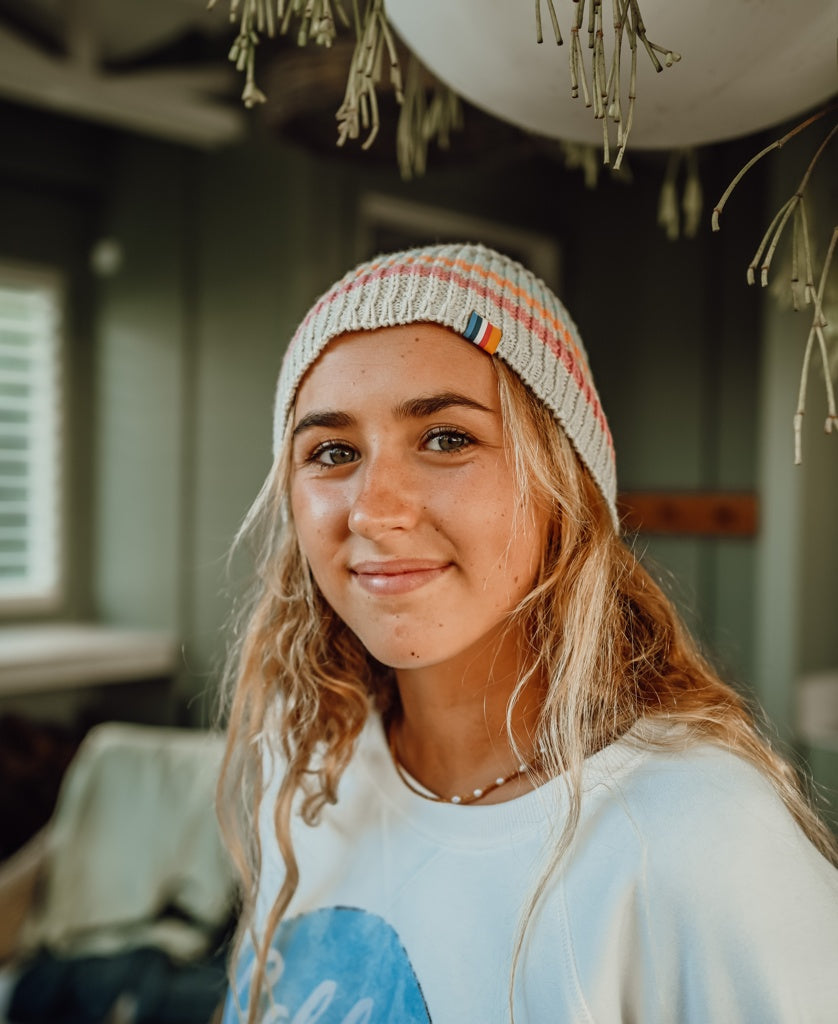 Young woman indoors wearing an Organic Cotton Beanie Thin Stripe | Oatmeal with natural light highlighting the casual surf-inspired style in a cozy setting.