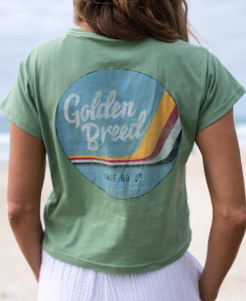 A person wears the Hattie Tee in Moss by Golden Breed, a soft cotton green cropped top featuring a vintage print and colorful back stripes, standing outdoors near the beach.