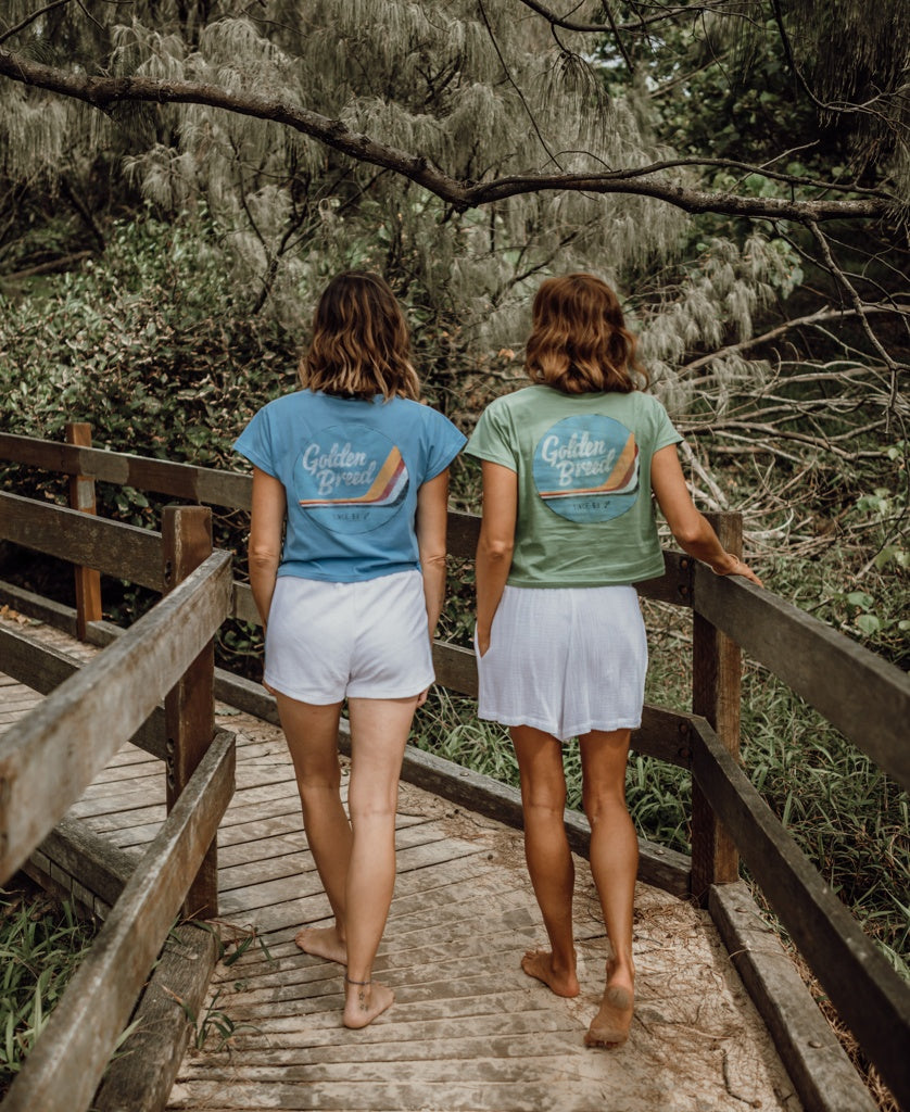 Two women with short brown hair walk side by side on a wooden path through greenery, wearing Golden Breeds Hattie Tee in Azure paired with white shorts or skirts.