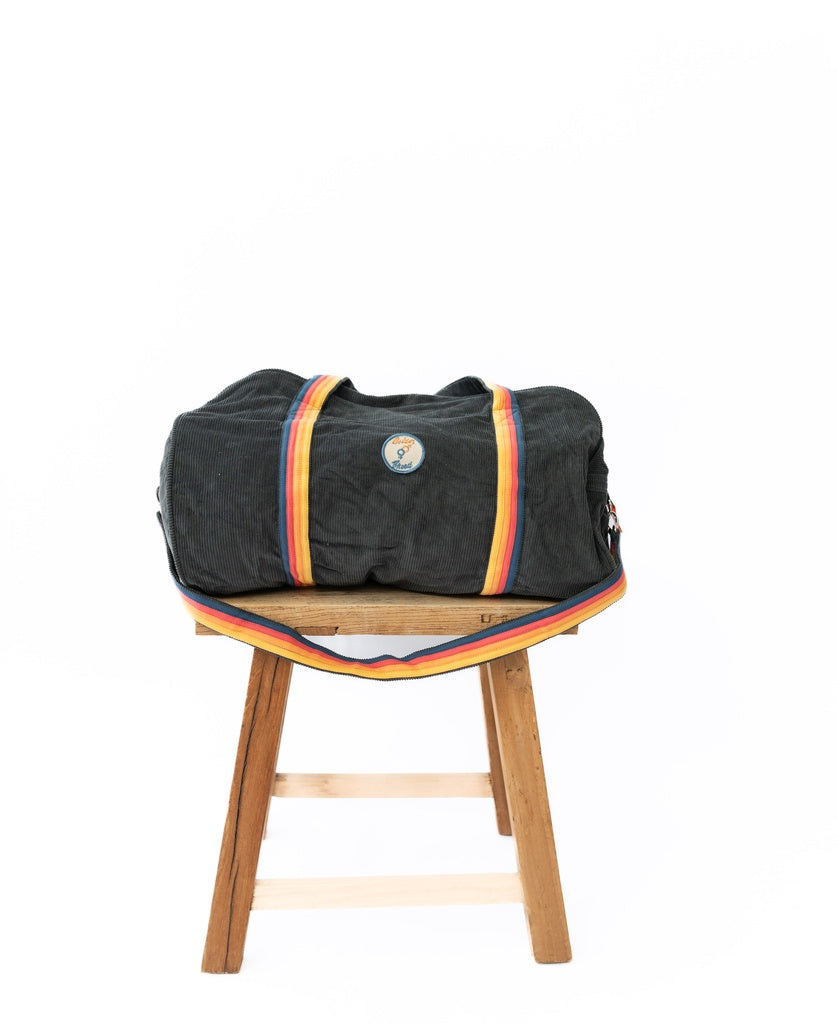 The Golden Breed Long Island Cord Roll Bag in Wash Black, featuring yellow and red striped cotton cord handles, is displayed on a wooden stool against a white background.