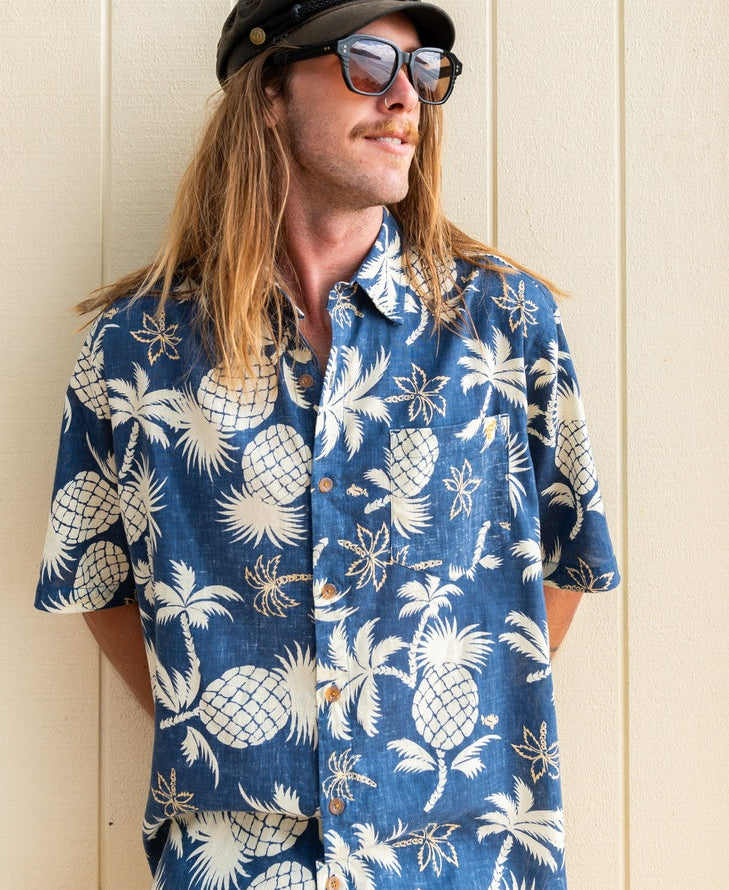 A person with long hair and a mustache wears the Golden Breed Pine-Palm Reverse Shirt in Vintage Navy, featuring a blue pine palm print, along with sunglasses and a black cap, standing against a beige wall.