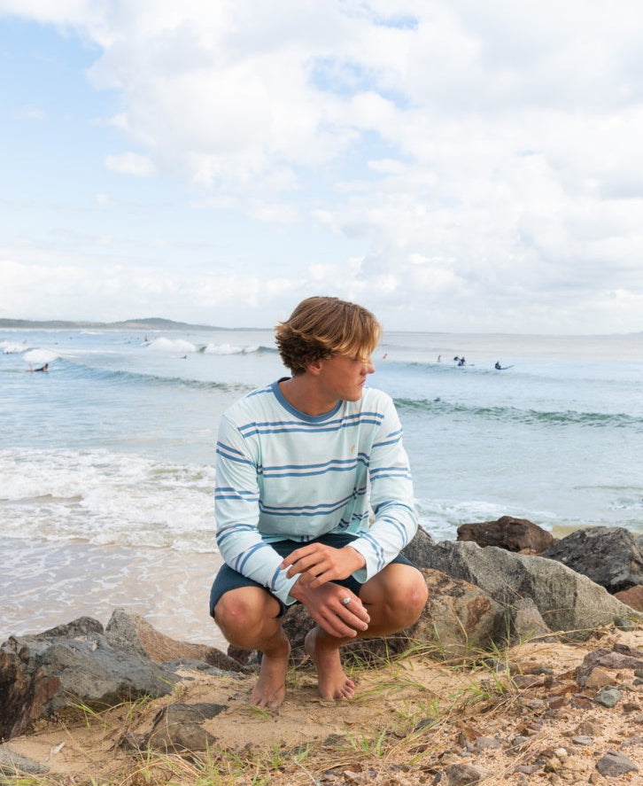 A young man in the Golden Breed Bay Stripe LS Tee | Ice and shorts crouches on a rocky beach, gazing sideways, channeling the Cali surf spirit with waves and surfers behind him.