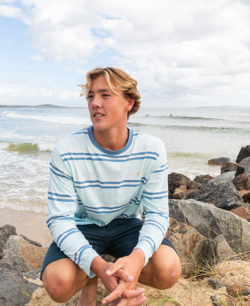 A young man with blond hair in the Golden Breed Bay Stripe LS Tee | Ice and shorts sits on rocks by the beach, waves crashing behind him, perfectly capturing the Cali surf vibe and beach lifestyle.