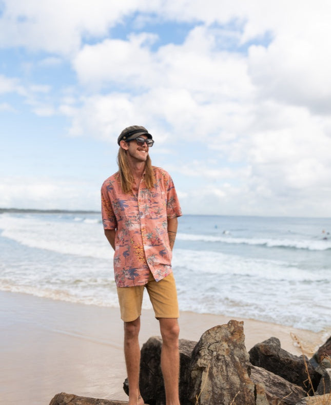 A man in a Golden Breed Flamingos Shirt | Rose, paired with shorts and a cap, stands barefoot on beach rocks as waves crash behind him under a cloudy sky.
