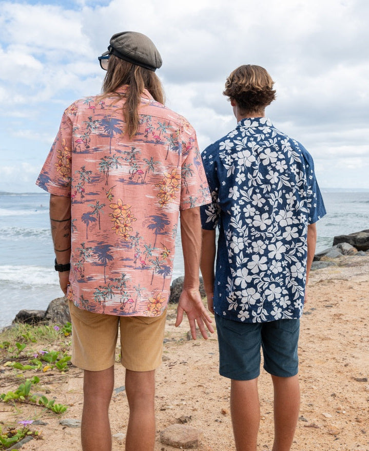 Two people stand side by side on sandy ground near rocks and greenery, facing the ocean while wearing Golden Breed’s Vomo Floral Shirt in Vintage Navy.