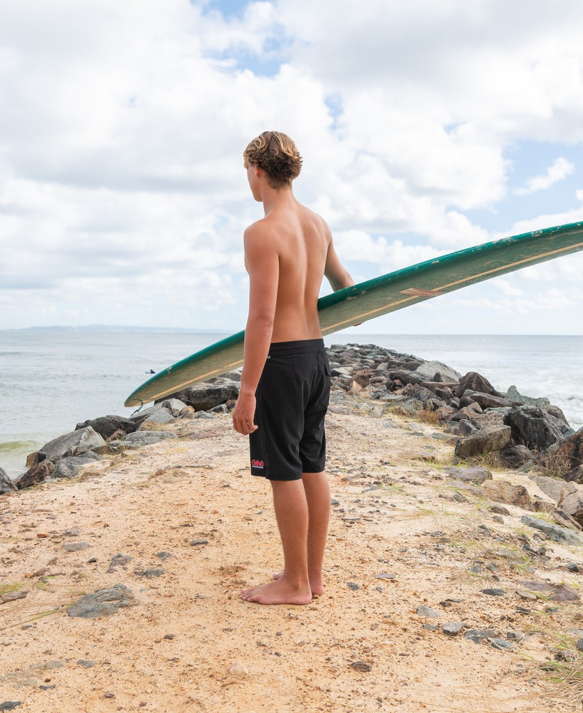 Barefoot on a sandy shore, a person gazes at the ocean with the Gordon & Smith G&S Boardie | Black—crafted from recycled polyester—tucked under one arm.