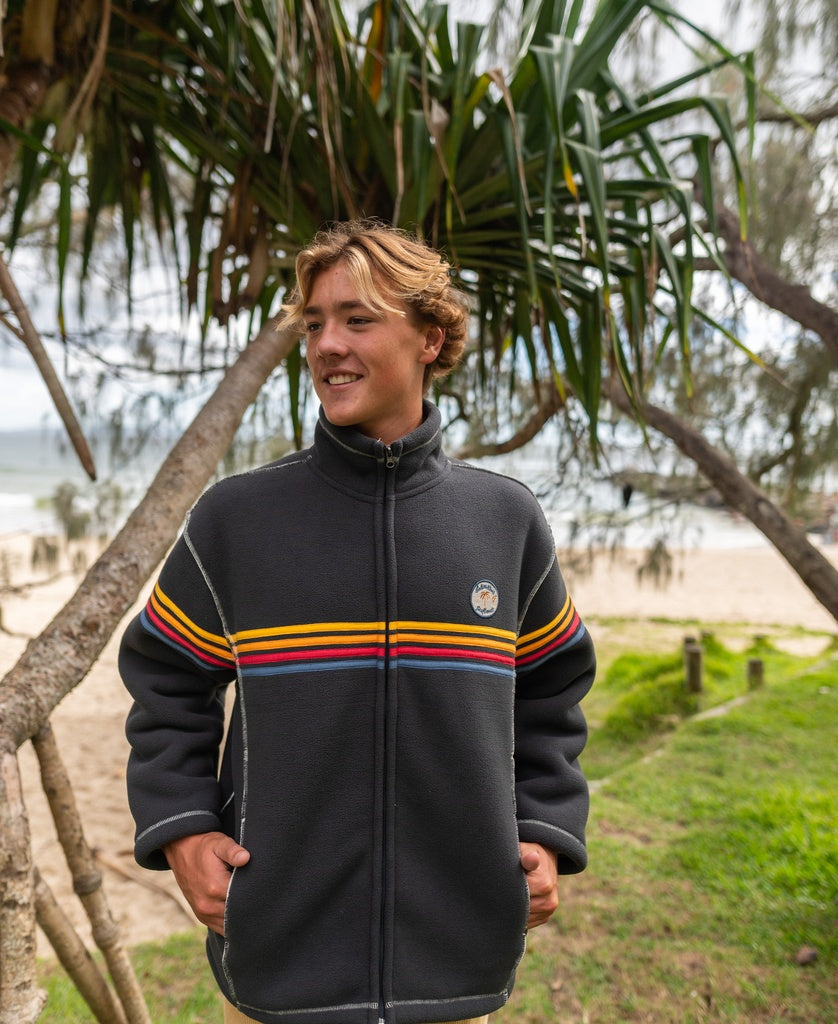 A young person with wavy blond hair smiles outdoors near a beach, wearing the Golden Breed Reef Bonded Sherpa Jacket in Wash Black, featuring yellow and red chest stripes and made from cozy polyester sherpa.